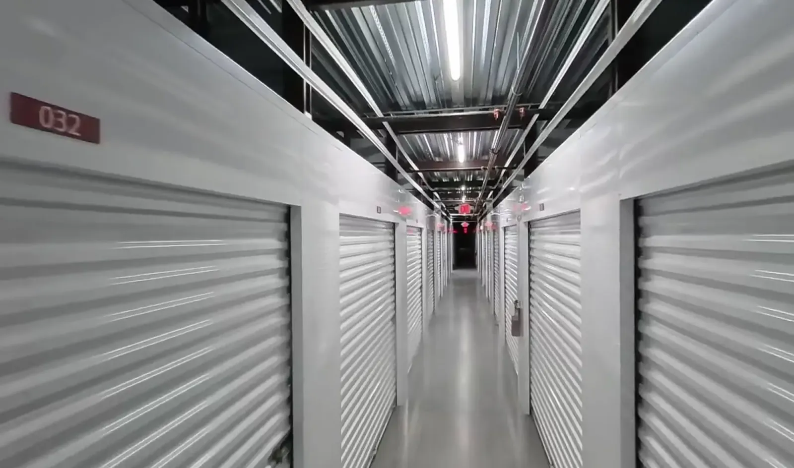 Indoor corridor lined with white roll-up unit doors under an exposed metal ceiling with overhead lighting